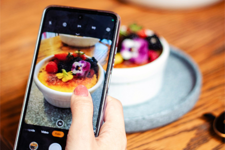 Hand holding smartphone capturing an image of a white ramekin filled with crème brûlée topped with edible flowers and berries, placed on a round blue plate on a wooden table.
