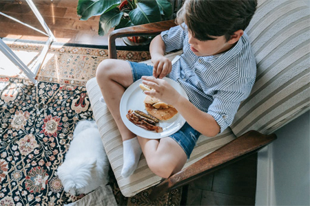 Photo of a pre-teen boy eating breakfast while sitting on a chair next to his cat