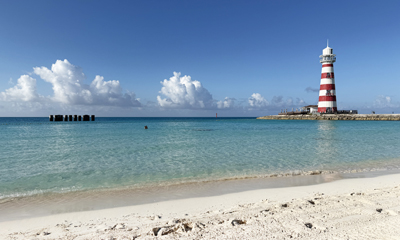 Close-up of the Ocean Cay lighthouse on a sunny day, with clear blue sky and calm sea gently lapping the white sandy beach.