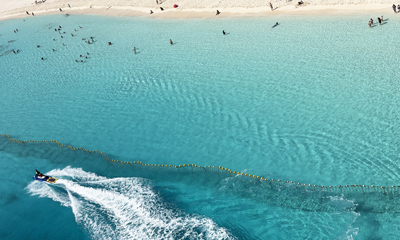 Aerial shot of a jet ski speeding across crystal-clear turquoise water near Ocean Cay, with swimmers visible in the water beyond a safety buoy line.